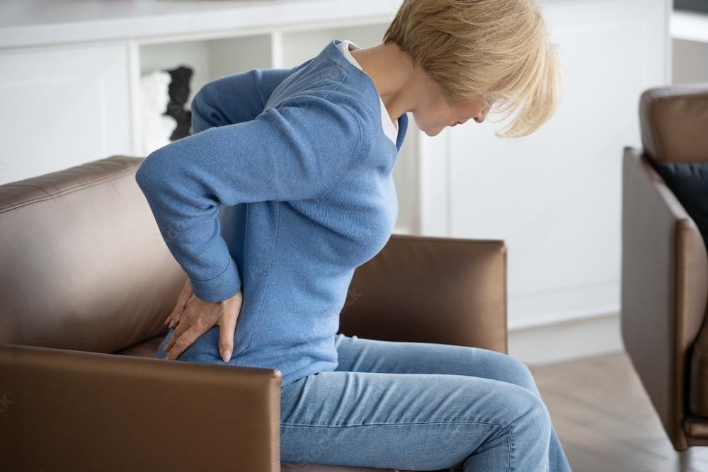 Middle aged woman feeling back pain, sitting on armchair