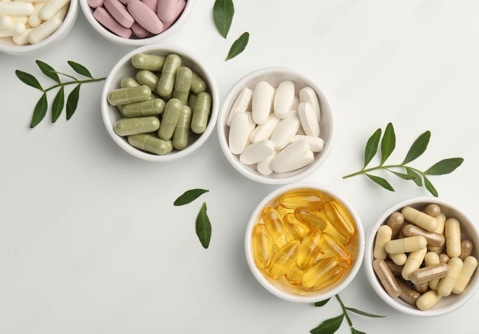 Different vitamin capsules in bowls and leaves on white background, flat lay. 